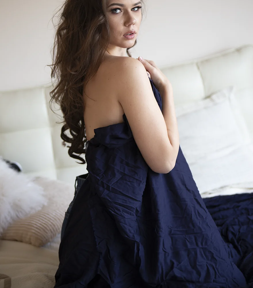 Woman in a wrinkled navy dress photographed naturally on a bed.