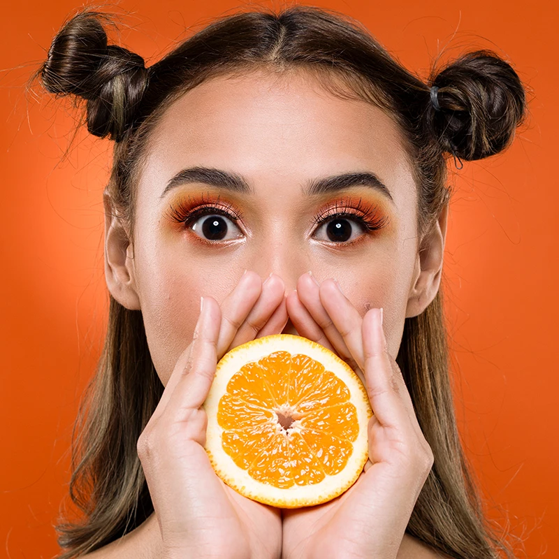 Unedited portrait of a woman holding an orange half in front of her face against an orange background.